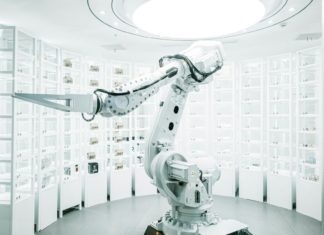 Vratky Zalanda v Německu a Polsku třídí roboti s umělou inteligencí Bílé průmyslové robotické rameno mezi regály s očíslovanými skladovacími boxy