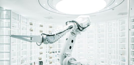 Vratky Zalanda v Německu a Polsku třídí roboti s umělou inteligencí Bílé průmyslové robotické rameno mezi regály s očíslovanými skladovacími boxy