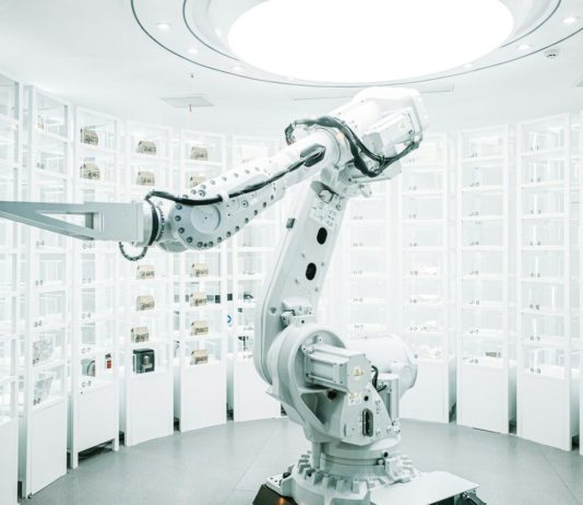 Vratky Zalanda v Německu a Polsku třídí roboti s umělou inteligencí Bílé průmyslové robotické rameno mezi regály s očíslovanými skladovacími boxy