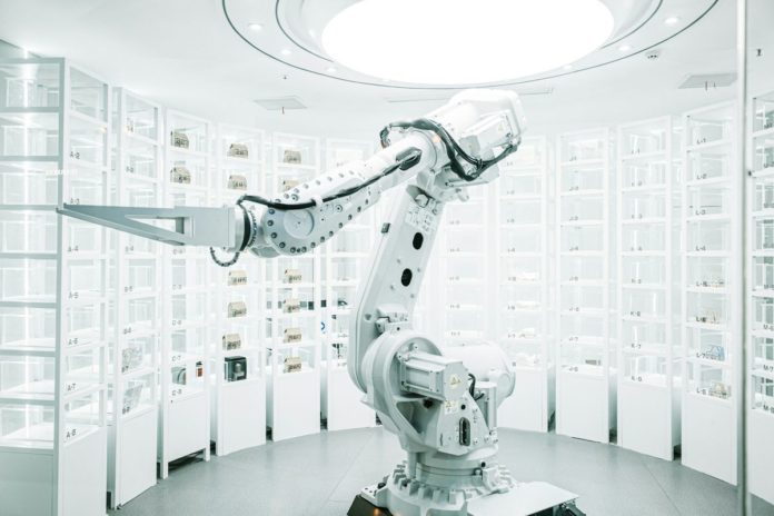 Vratky Zalanda v Německu a Polsku třídí roboti s umělou inteligencí Bílé průmyslové robotické rameno mezi regály s očíslovanými skladovacími boxy
